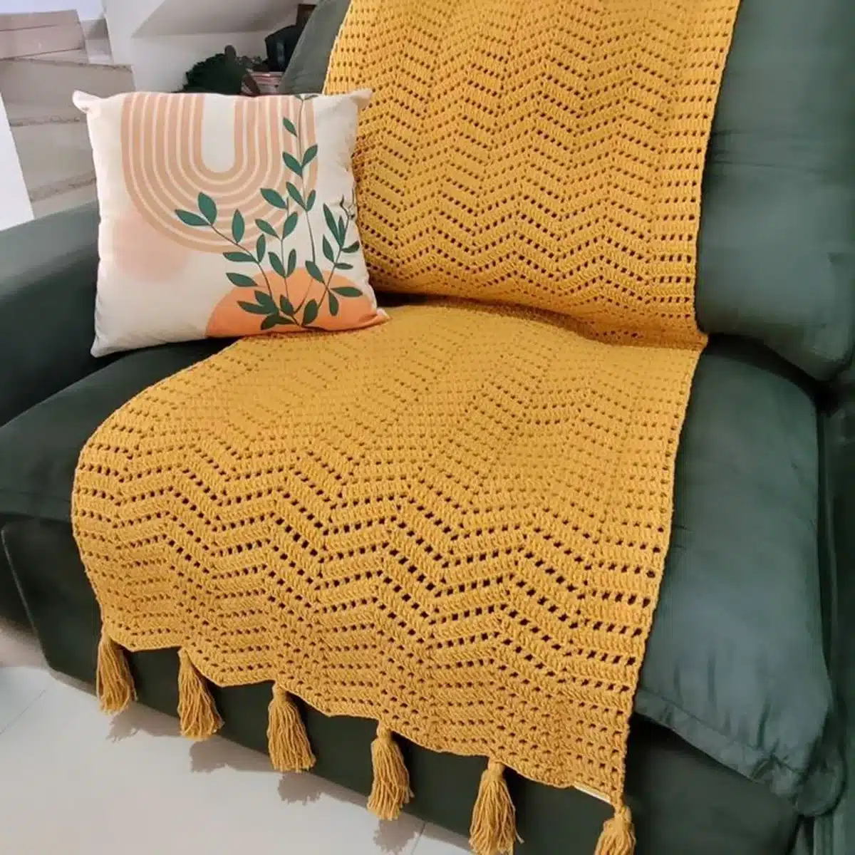 Manta de crochê amarela com franjas sobre sofá verde e almofada decorativa com estampa moderna.