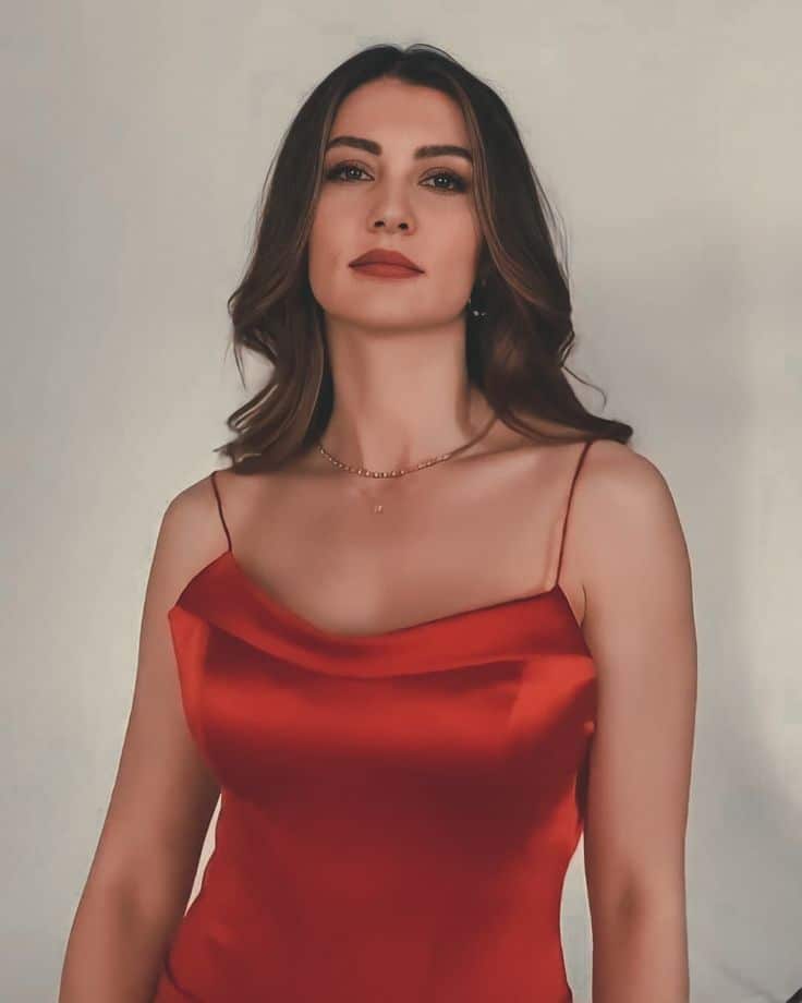 Atriz turca Burcu Özberk usando vestido vermelho, olhando diretamente para a câmera, com cabelo solto e maquiagem suave.