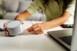 Pessoa ajustando um alto-falante inteligente em uma mesa, próximo a um laptop.