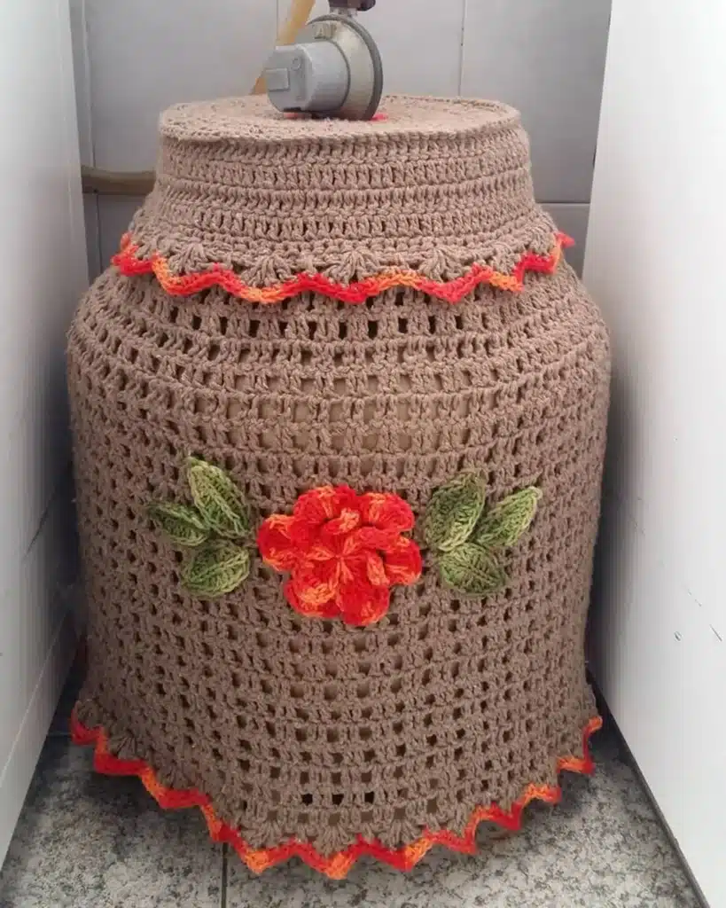 Capa de crochê marrom com bordas em tons de laranja e flor vermelha decorativa para botijão de gás.