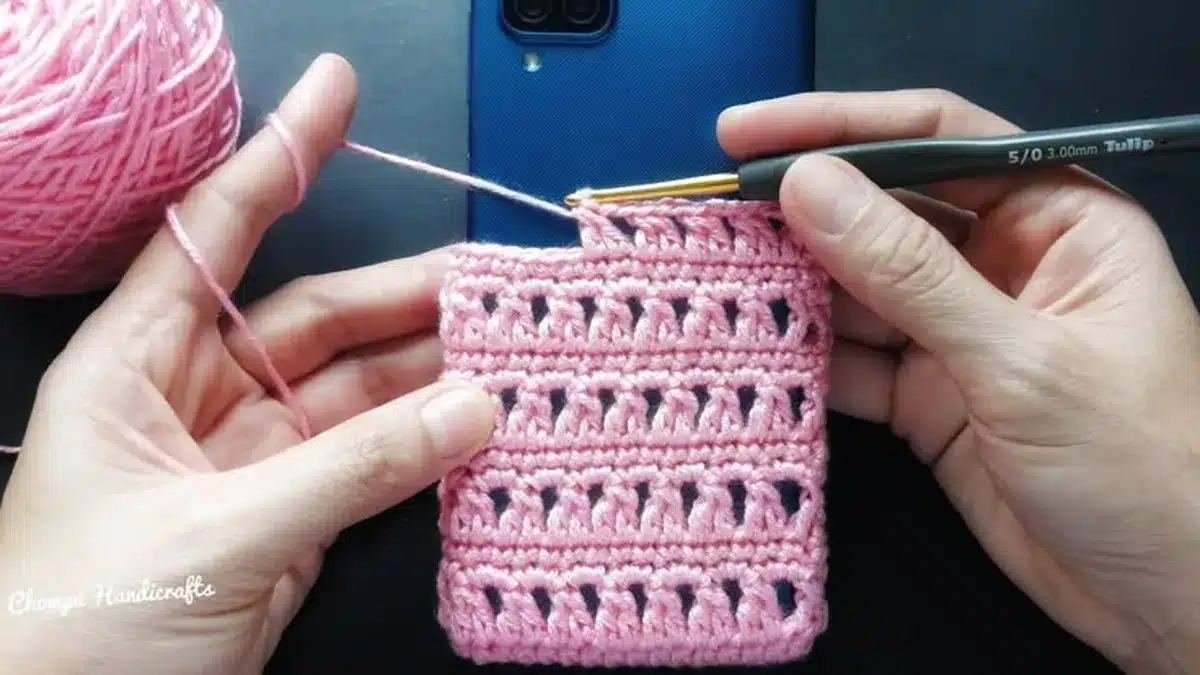 Mãos crochetem uma capinha rosa para celular, com dispositivo azul ao fundo.