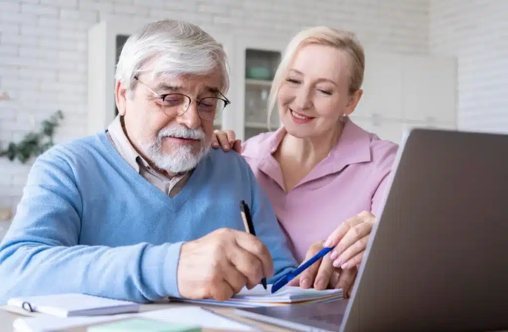 Casal idoso feliz planejando a aposentadoria juntos, com papéis e laptop à frente.