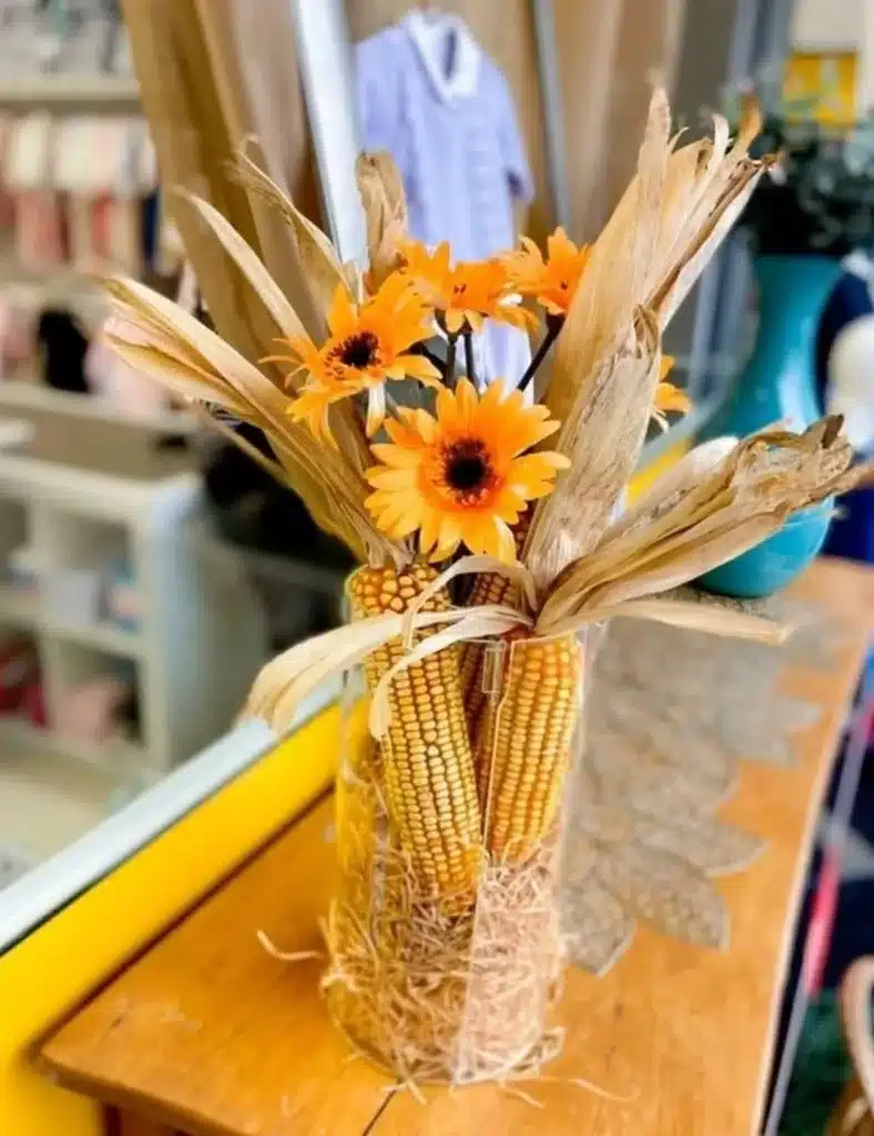 Arranjo decorativo de centro de mesa com espigas de milho seco e flores amarelas em jarra de vidro