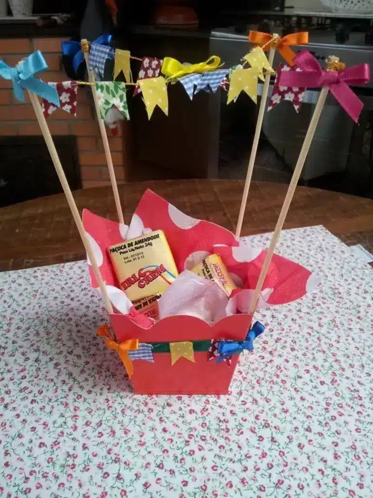 Centro de mesa decorativo com bandeirinhas coloridas e paçoca em embalagem dentro de caixa vermelha
