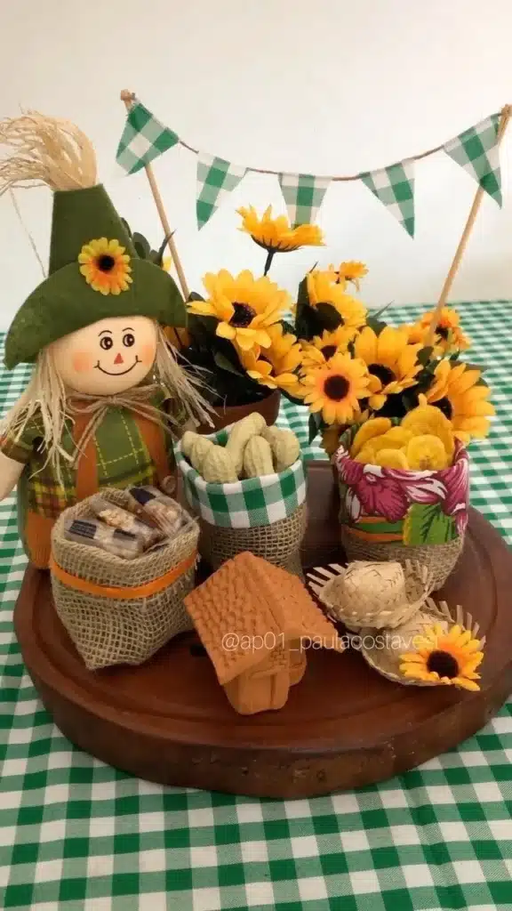 Centro de mesa decorativo com boneco caipira, flores amarelas, amendoim e salgadinhos em sacos de juta