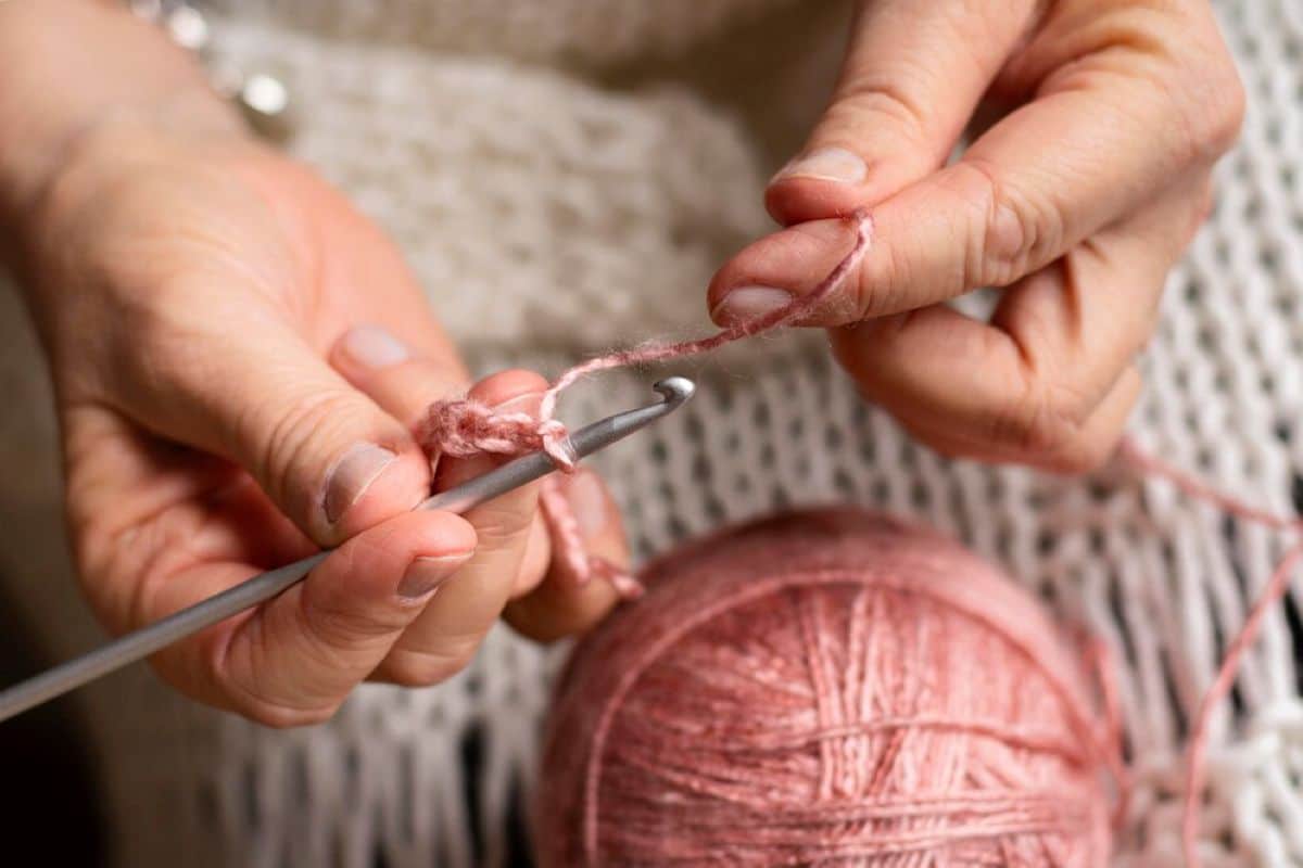 Mãos femininas fazendo crochê com fio rosa, representando o processo artesanal de criação de peças para cozinha