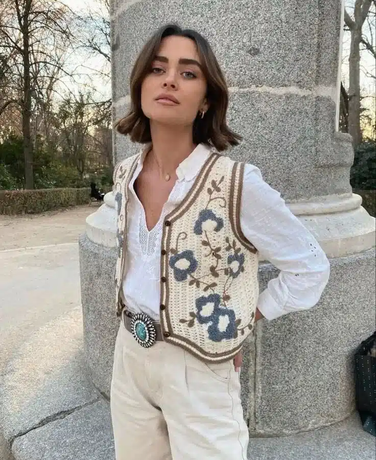 Mulher usando colete de crochê com flores em tons de azul e bege, com uma camisa branca e calças claras, posando em ambiente ao ar livre.