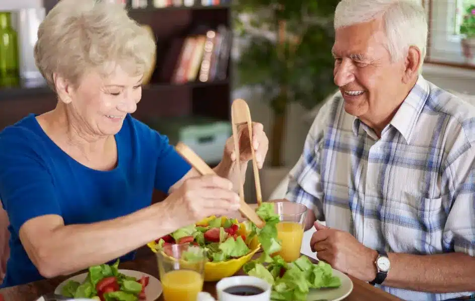 Casal idoso feliz compartilhando uma refeição saudável com salada fresca e suco de laranja.