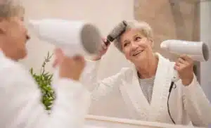 Mulher idosa sorridente secando os cabelos com secador e escova em frente ao espelho