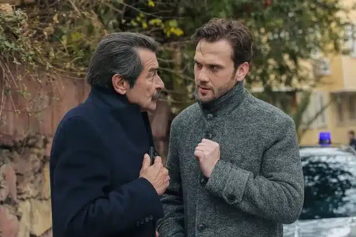 Dois homens conversando de forma intensa em uma rua arborizada, vestidos com casacos de inverno.