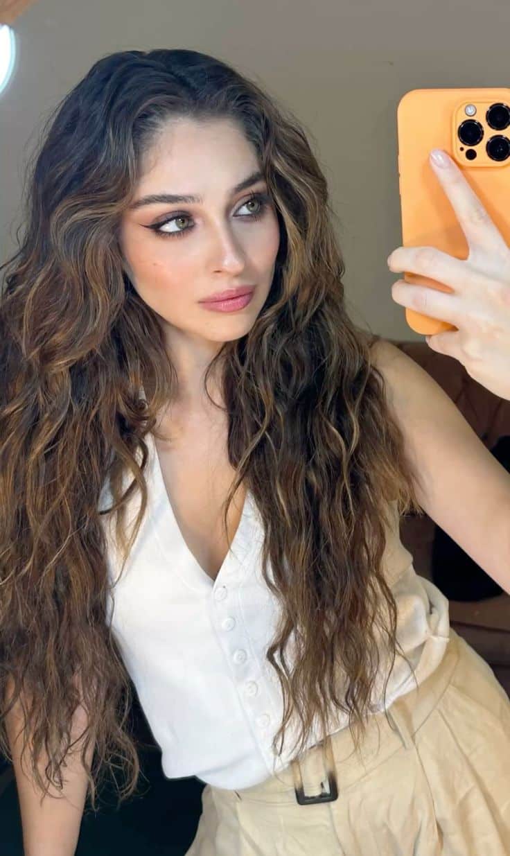 Atriz turca Cemre Baysel tirando selfie, com cabelos longos e ondulados, usando blusa branca e calça bege, em ambiente interno.