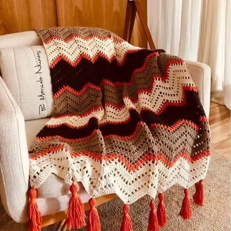  Manta colorida de crochê com franjas vermelhas, dobrada casualmente sobre um sofá bege em uma sala aconchegante.