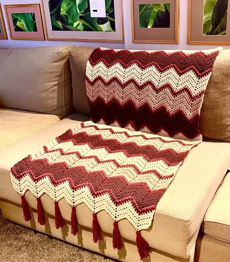 Manta de crochê vermelha, creme e bordô com franjas, posicionada sobre um sofá bege em ambiente aconchegante.