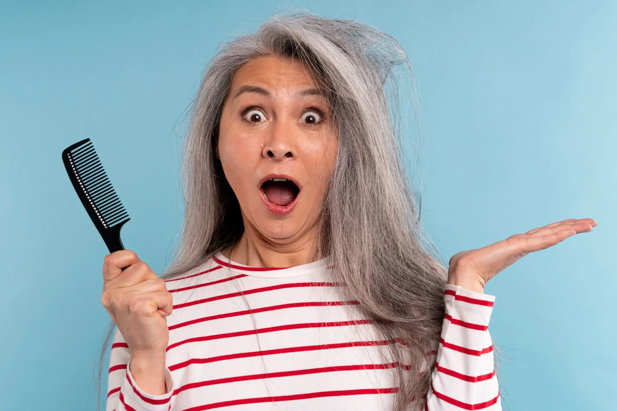 Mulher madura segurando um pente, surpresa com o cabelo bagunçado e ressecado sobre fundo azul.