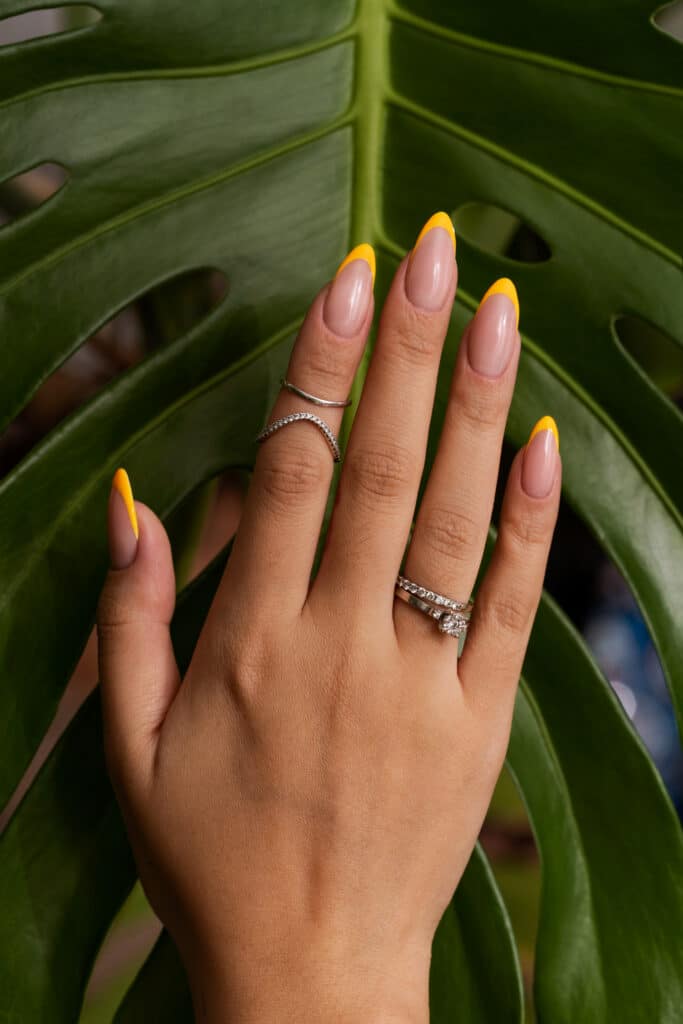 Unhas com francesinha moderna em amarelo vibrante com fundo de folhas verdes tropicais.