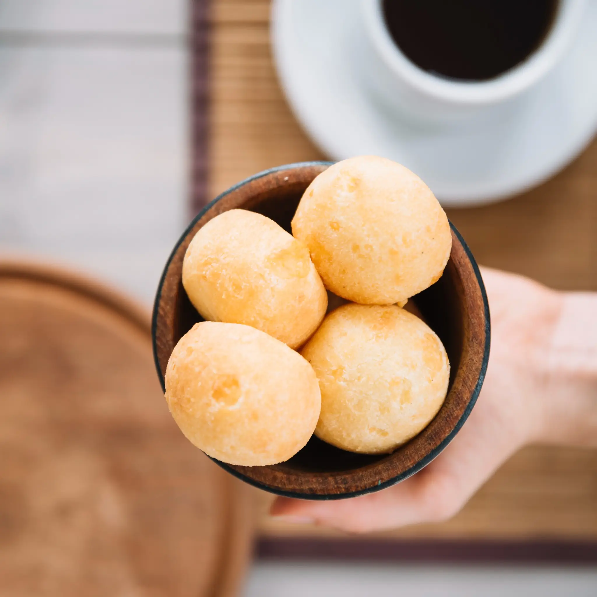 Pão de queijo mineiro sem ovos servido em tigela de madeira, com café ao fundo