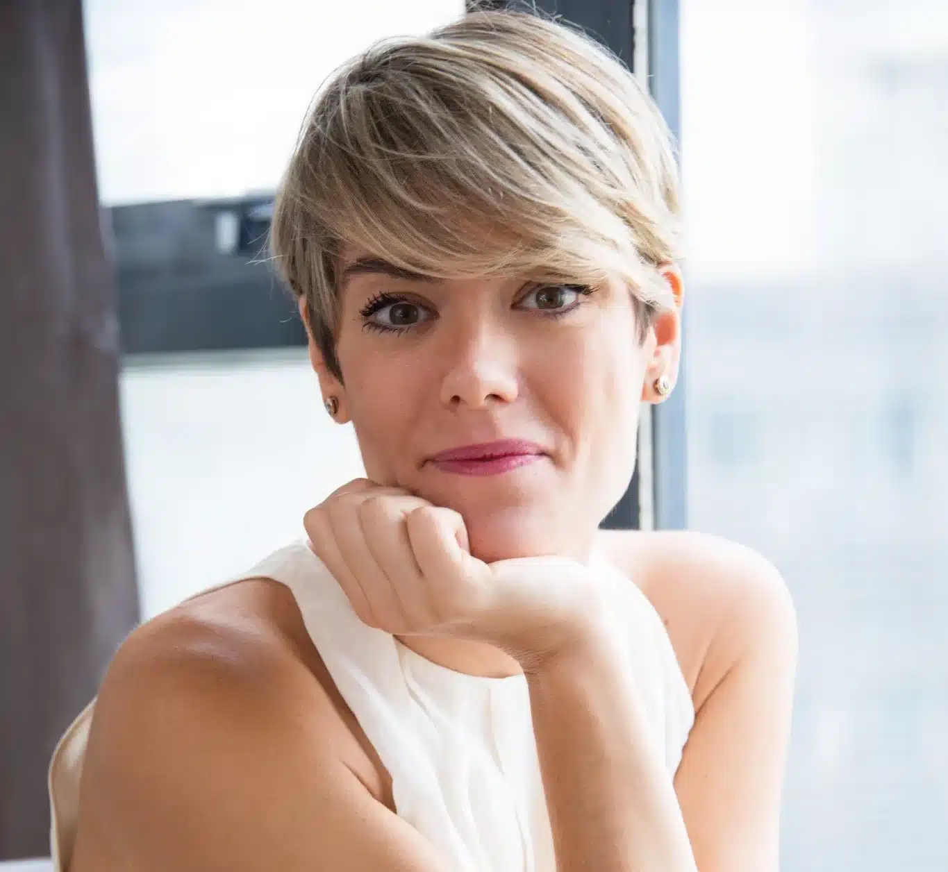 Mulher com corte pixie moderno, sorrindo e inclinando o rosto sobre a mão, mostrando estilo e confiança.