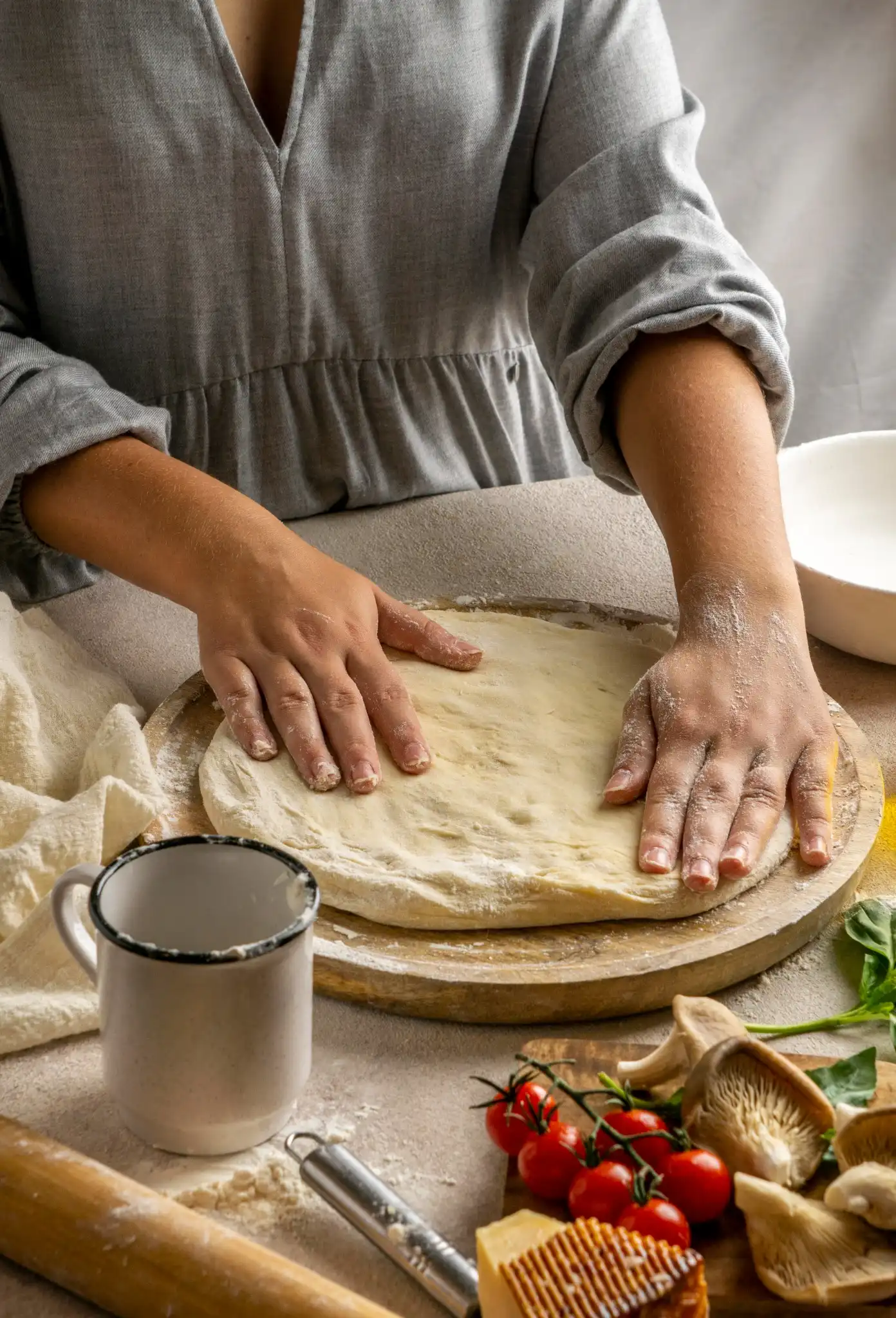 Mão feminina esticando a massa de pizza caseira sobre a bancada, com ingredientes ao fundo.