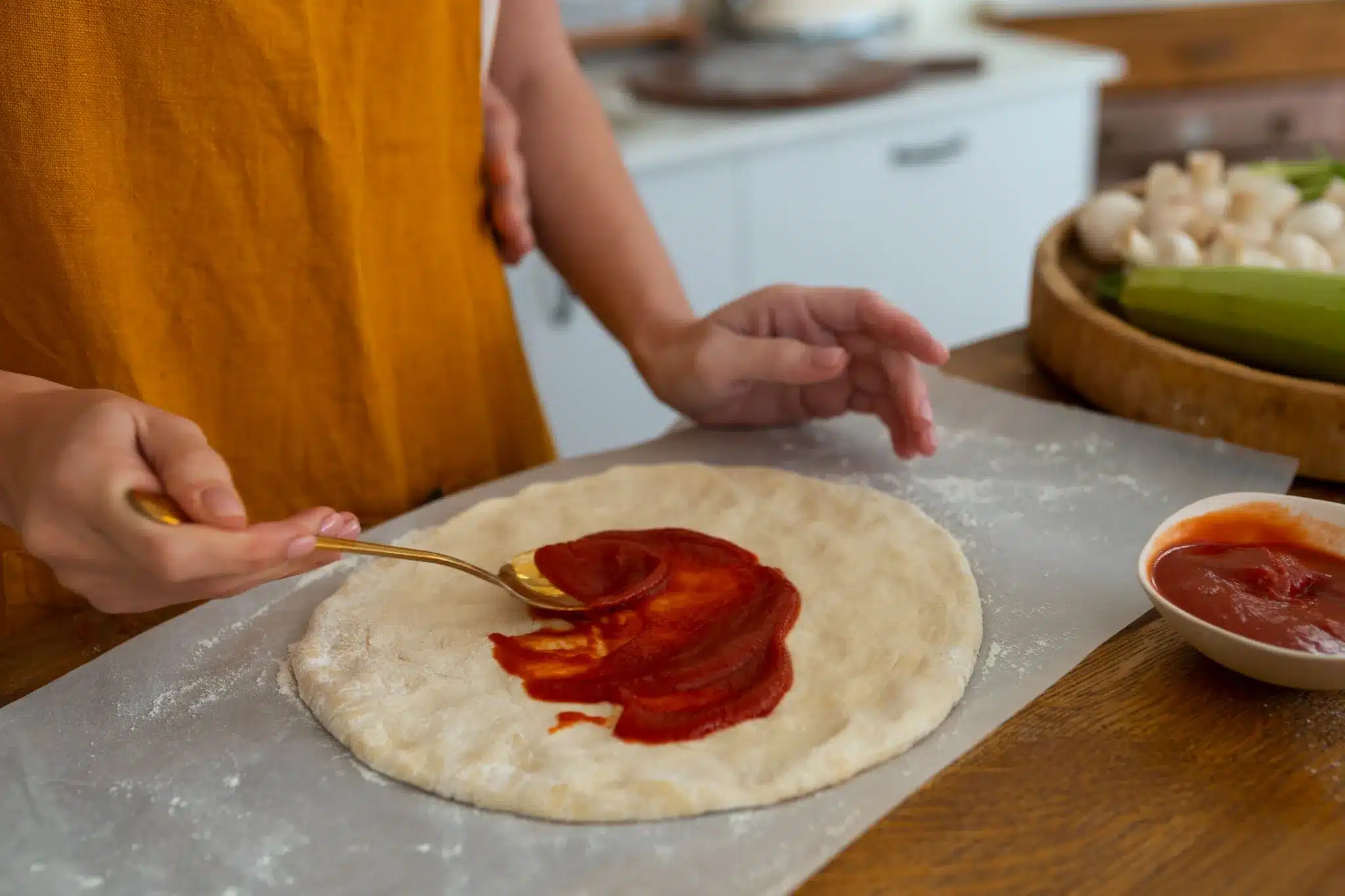 Pessoa espalhando molho de tomate sobre a massa da pizza caseira com uma colher dourada.