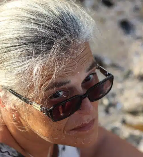 Mulher madura de óculos escuros com cabelos grisalhos exposta ao sol, representando os danos solares aos fios.