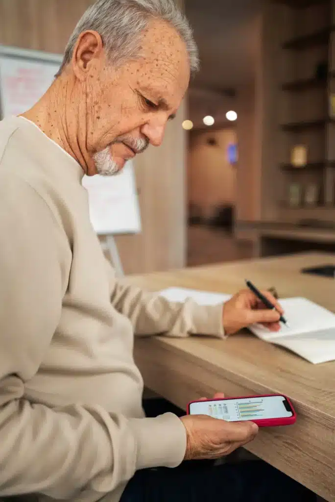  Homem idoso analisando gráficos financeiros no celular e anotando em caderno.