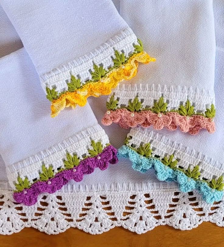 Detalhe de barrados de crochê coloridos em toalhas brancas, com flores em amarelo, rosa, roxo e azul.