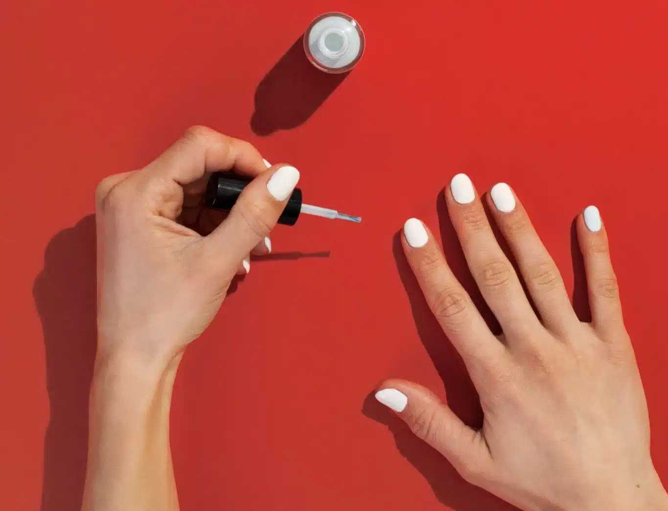 Mãos femininas esmaltando unhas brancas sobre fundo vermelho