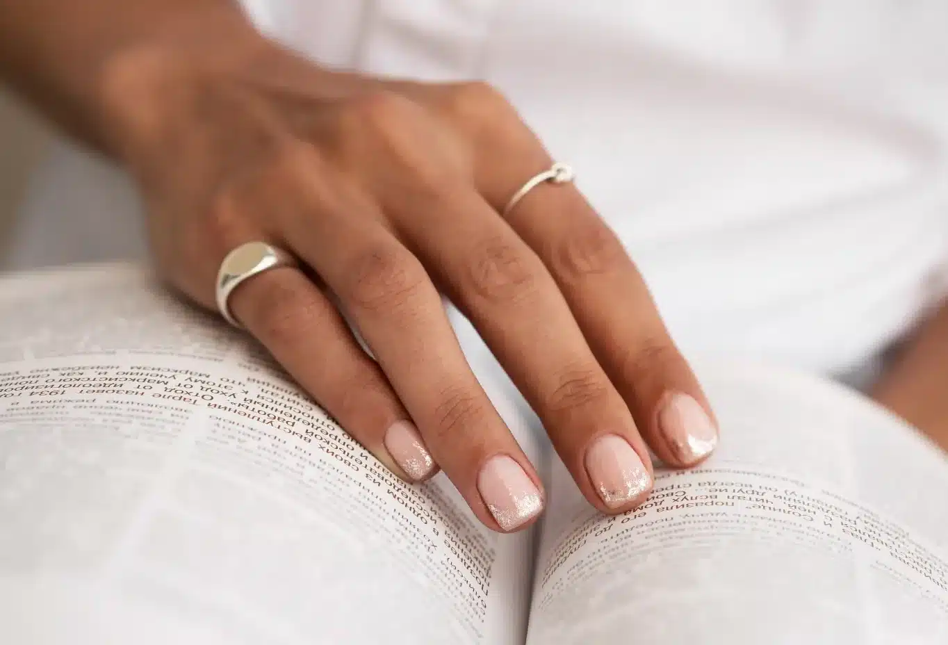 Mão feminina com francesinha delicada em tom nude e glitter, segurando um livro aberto.