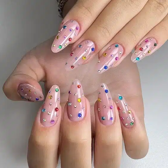 Unhas decoradas com base transparente, cobertas por pedrinhas coloridas e brilhos que criam um efeito delicado e festivo.