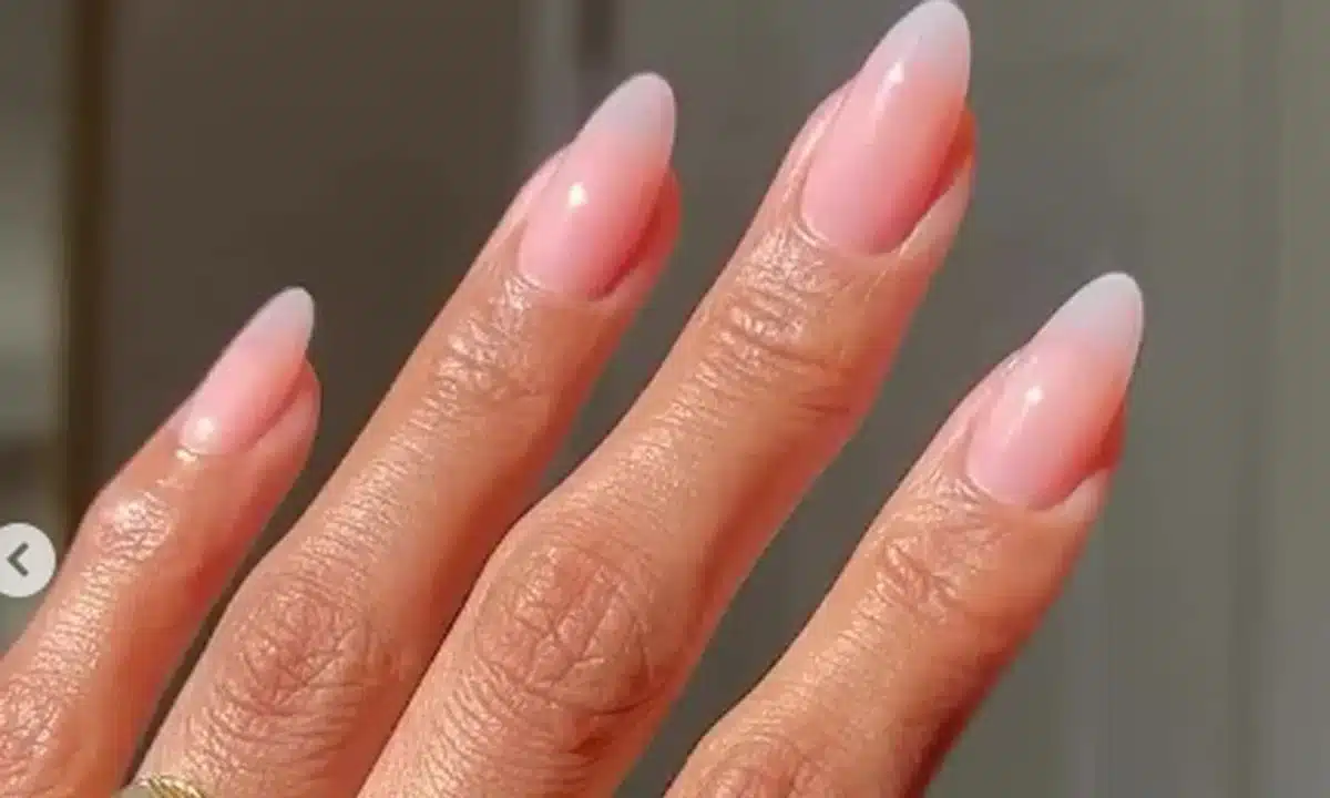 Unhas com esmalte em degradê rosa suave, formato amendoado, adornadas com anéis dourados minimalistas.
