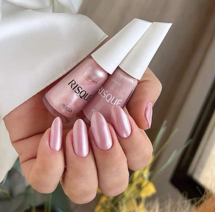 Mão segurando dois esmaltes Risqué em tons rosados com unhas pintadas no estilo cintilante