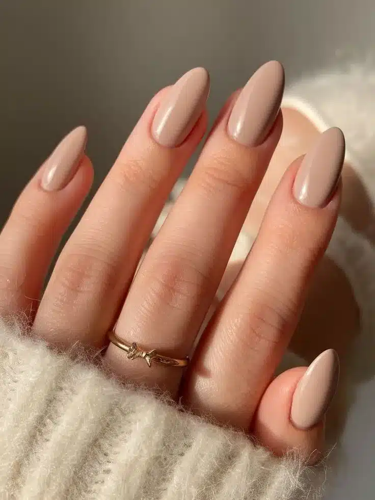 Unhas em tom bege amanteigado, com acabamento liso e anel dourado delicado.