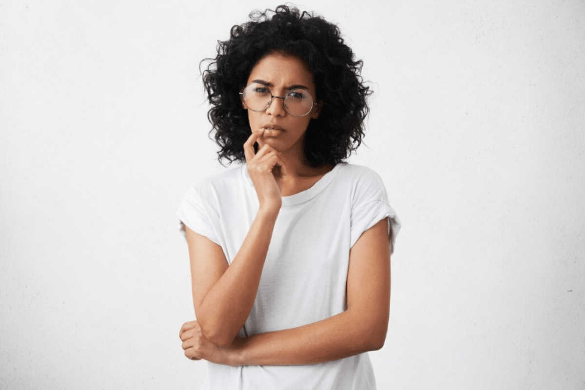 Pessoa pensativa com óculos, vestindo camiseta branca, em fundo neutro.