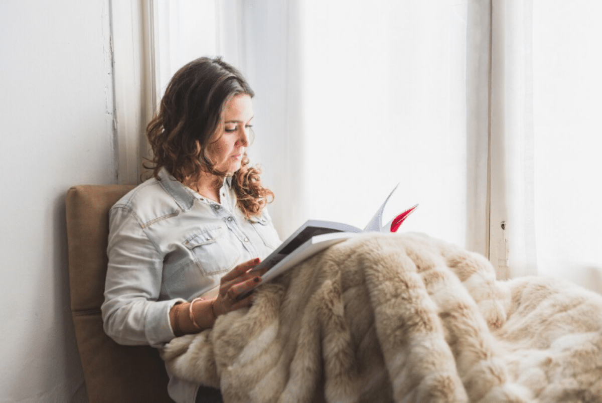 Mulher lendo um livro enquanto está sentada perto da janela com uma manta aconchegante