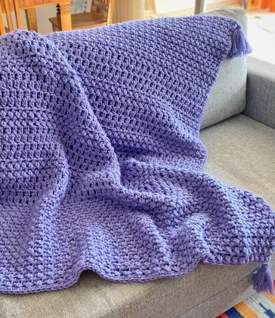 Manta de crochê na cor lavanda com pontos largos e acabamento com tassel