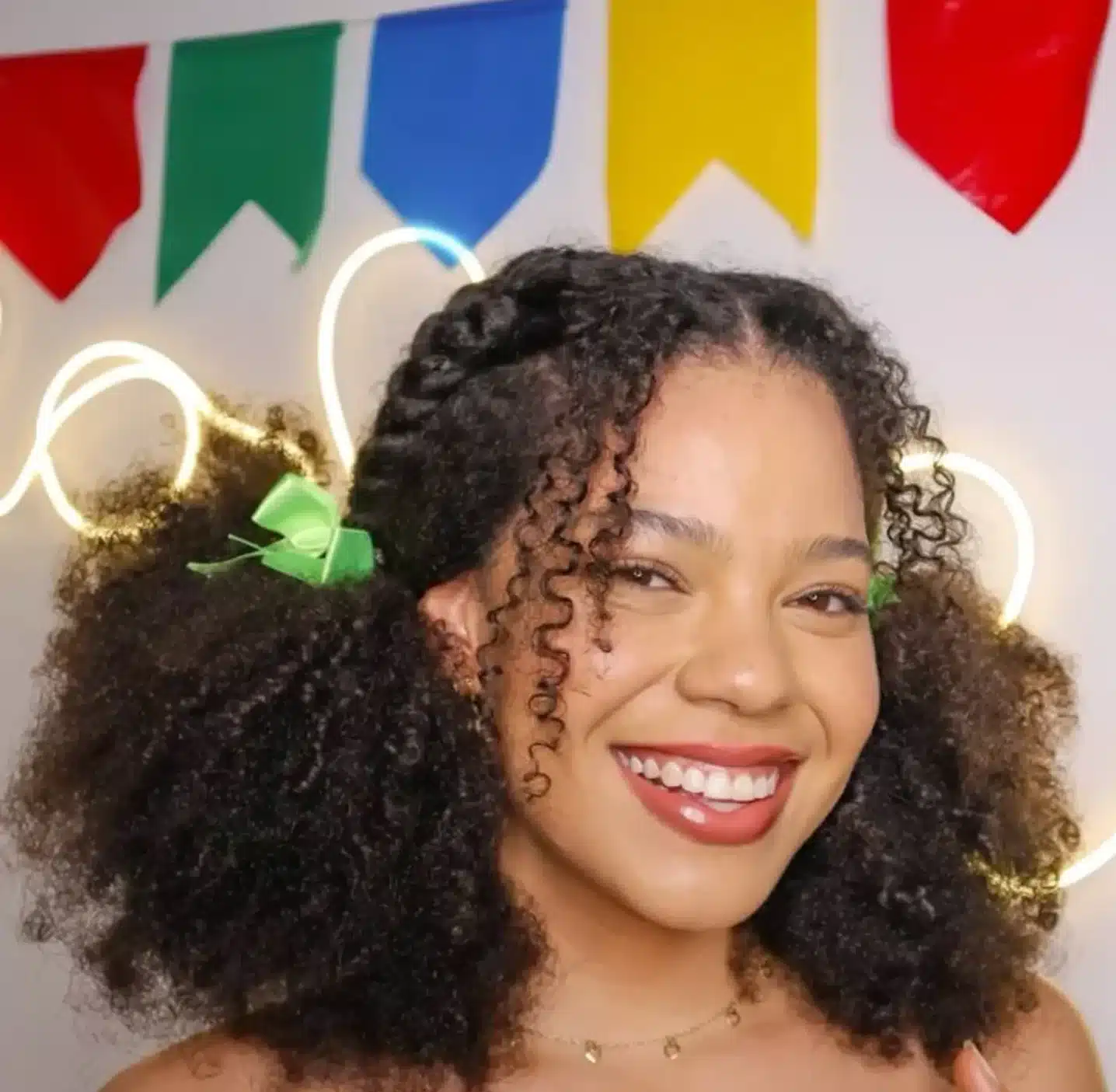 Mulher jovem com cabelos cacheados e laços verdes, sorrindo em ambiente colorido com bandeirinhas juninas no fundo.