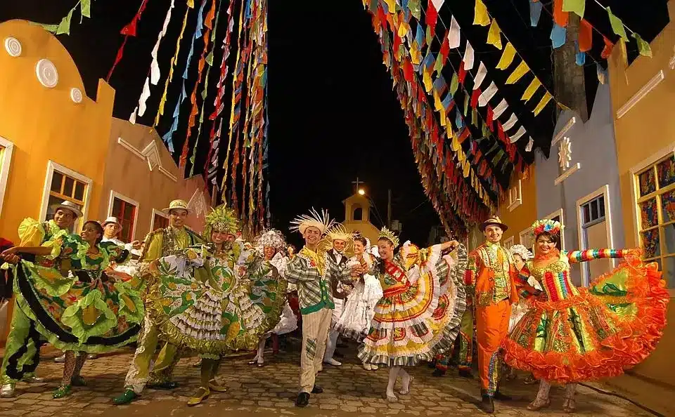Grupo de pessoas dançando quadrilha com trajes típicos de festa junina sob bandeirinhas coloridas