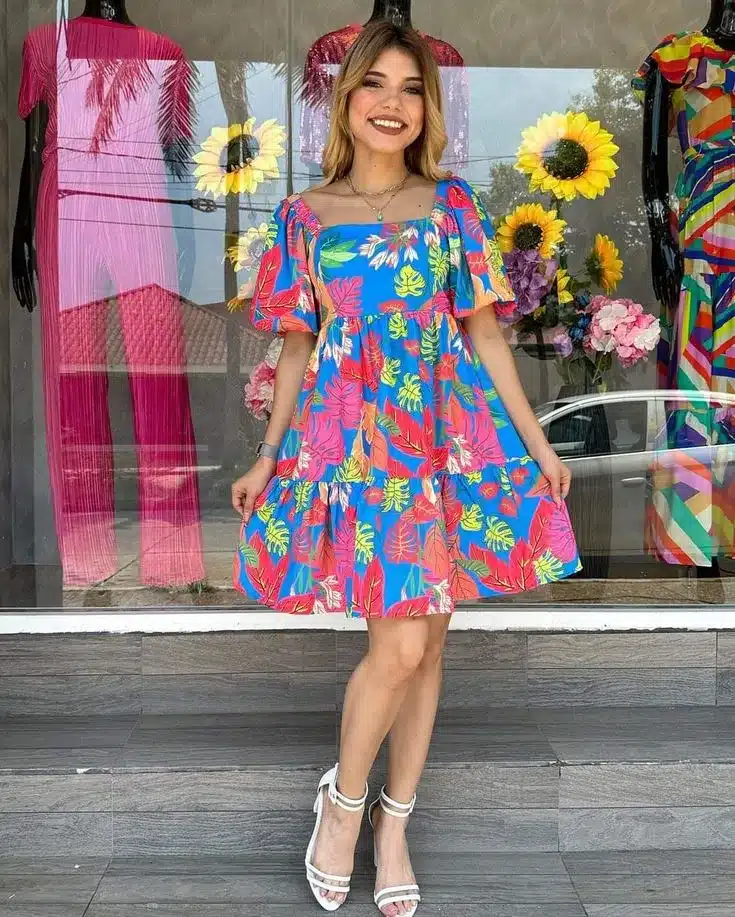 Modelo usando um vestido floral vibrante de cores vivas, perfeito para ocasiões descontraídas e alegres, com flores e folhas em tons de azul, vermelho e verde.