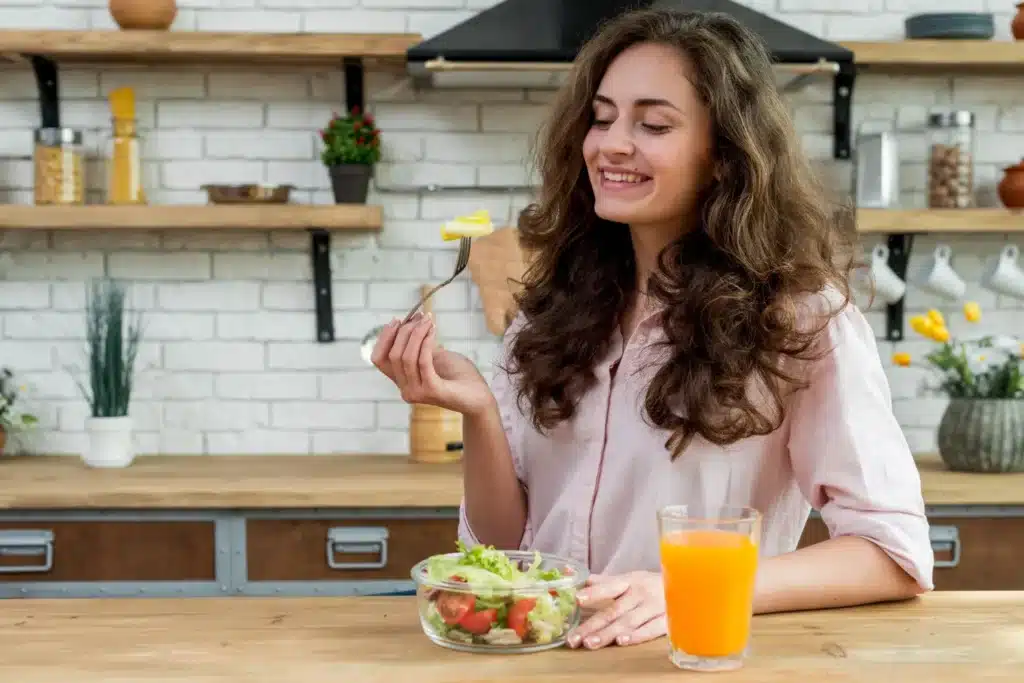 Mulher morena com cabelo cacheado comendo uma salada, com um copo de suco ao lado, em ambiente de cozinha.