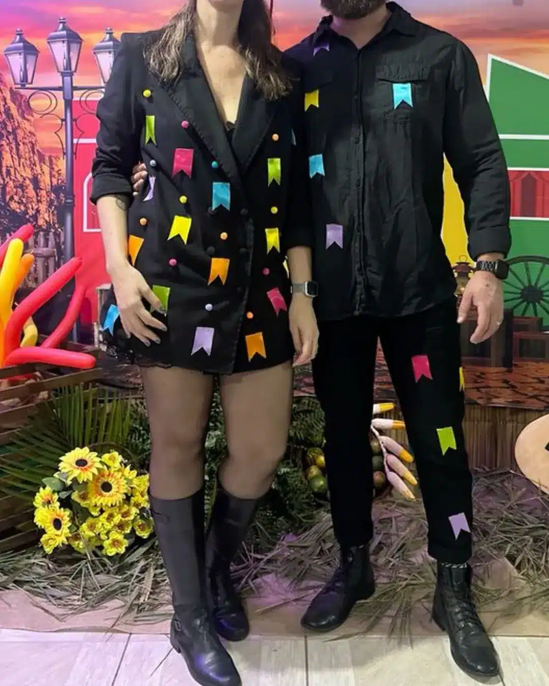 Casal vestindo roupas pretas decoradas com bandeirinhas coloridas em festa temática, cenário com flores e decoração vibrante ao fundo.