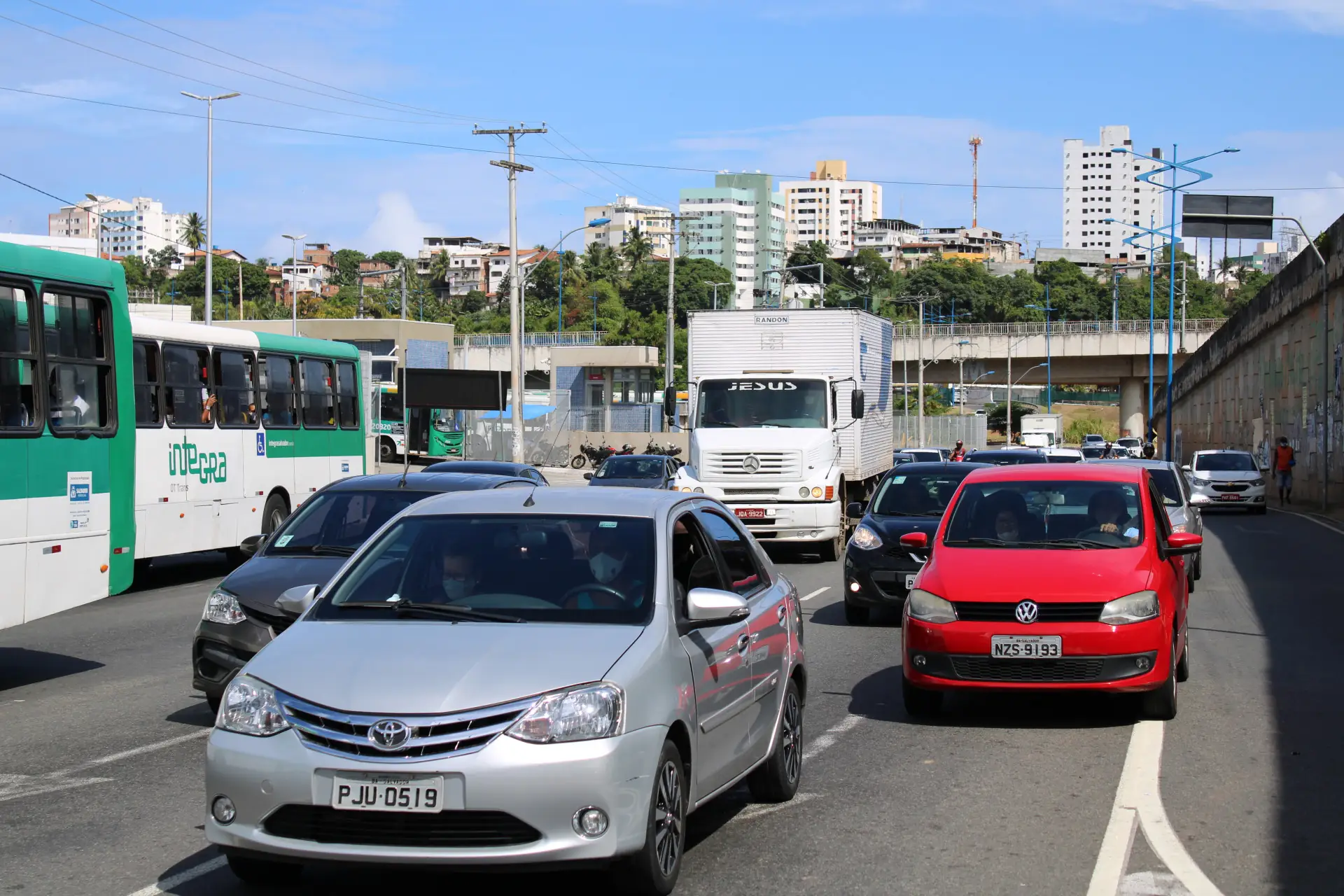 Trânsito congestionado em avenida urbana durante o dia, com carros, ônibus e caminhões parados.