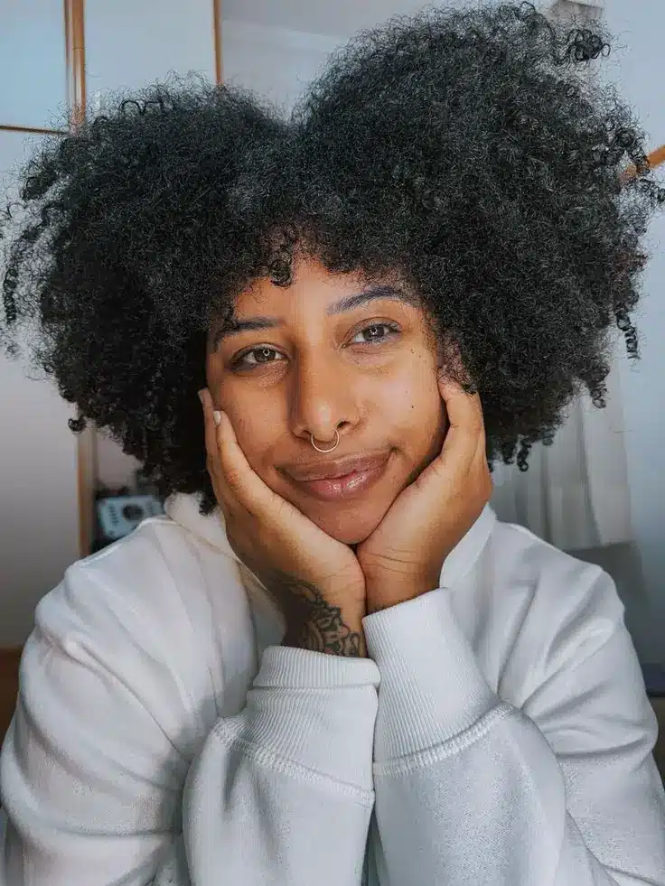 mulher negra com cabelo crespo em corte coração sorrindo com as mãos no rosto