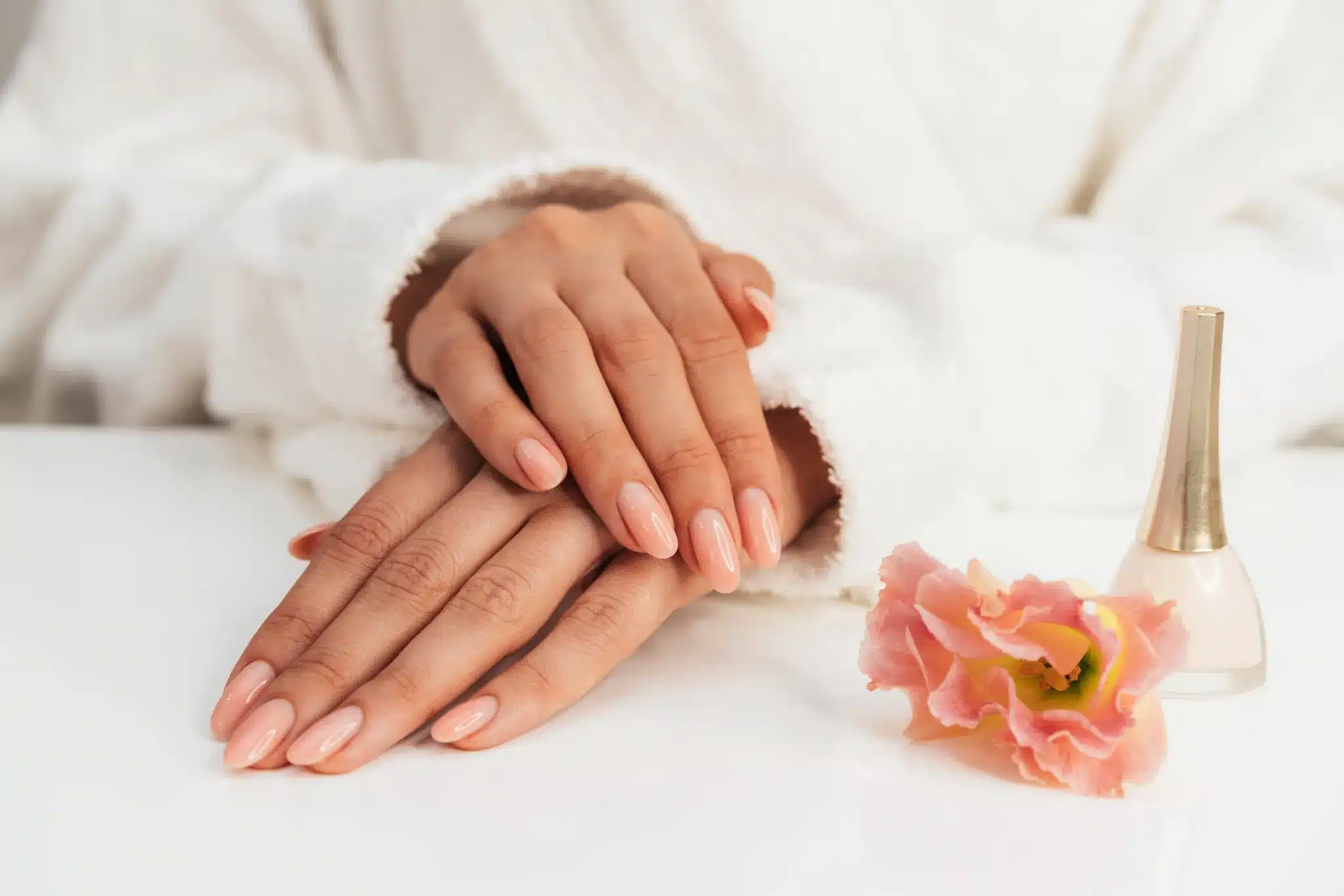 Mãos femininas com unhas bem feitas ao lado de esmalte e flor decorativa