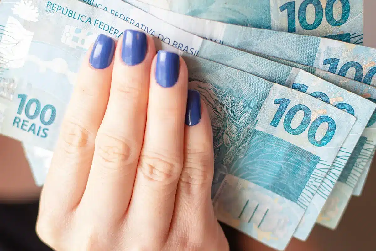 Notas de 100 reais sendo seguradas por uma mão com unhas esmaltadas.
