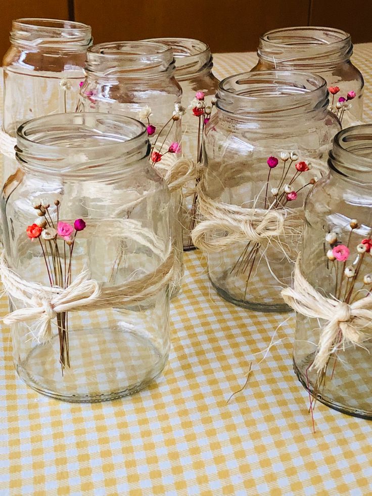 Potes de vidro decorados com flores secas e laços de barbante, usados como centro de mesa para festas.