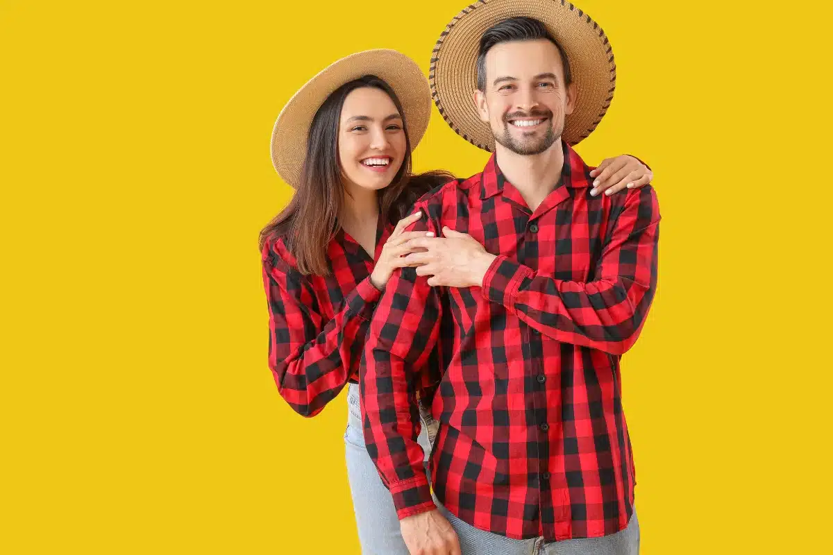 Casal sorridente vestindo camisas xadrez vermelhas e chapéus de palha, posando em fundo amarelo.