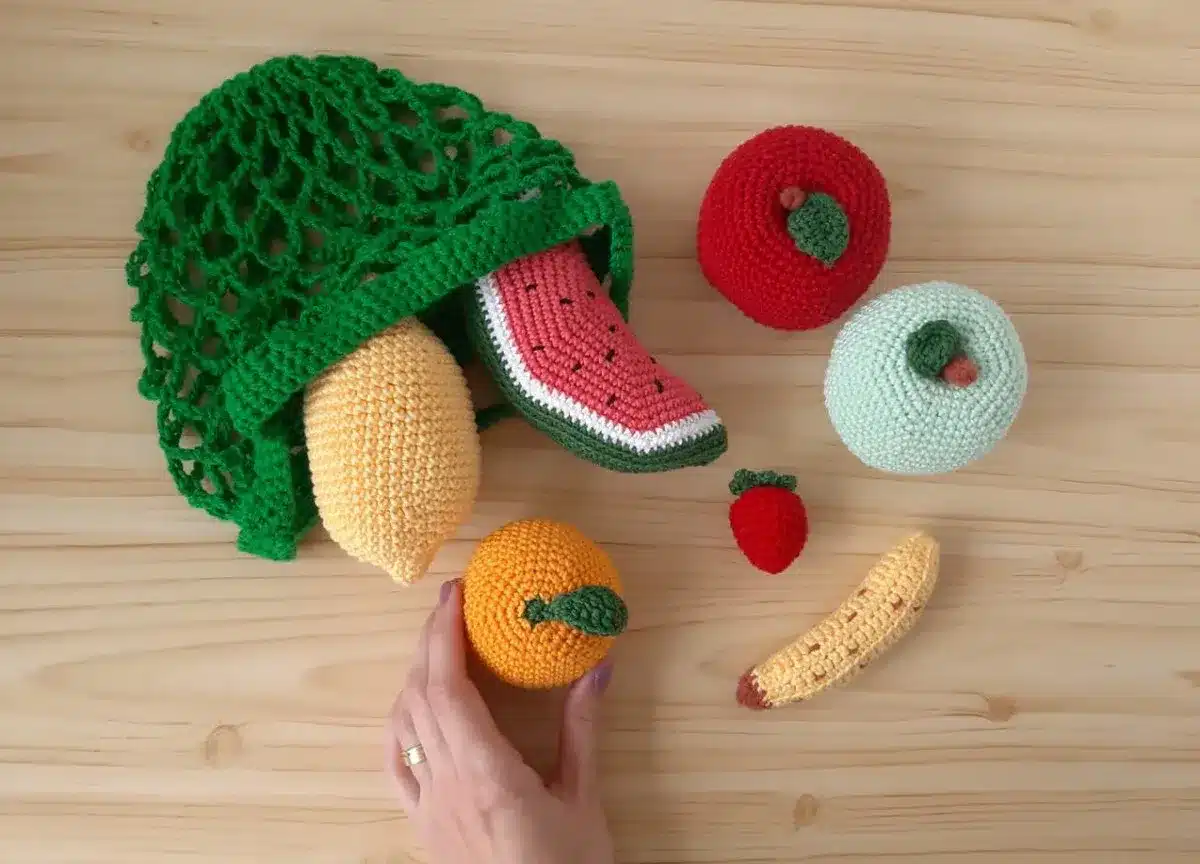 Amigurumis de frutas crochê, incluindo limão, melancia, maçã, morango e banana, em uma rede verde.