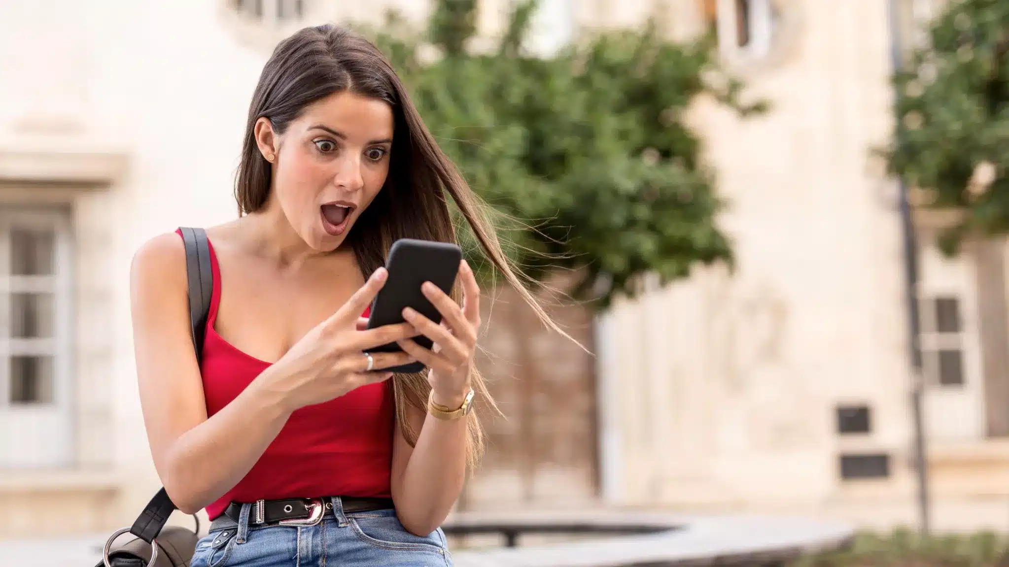 Mulher jovem surpresa ao ler mensagem de texto no celular, em ambiente externo.