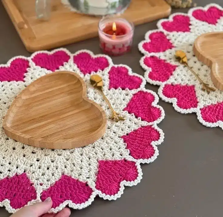 Sousplat de crochê com corações rosas e beiges, acompanhados de pratos de bambu e vela aromática.