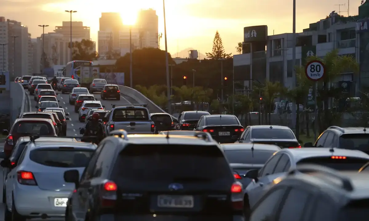 Trânsito intenso ao entardecer em avenida movimentada, com diversos carros parados e sinal vermelho ao fundo.